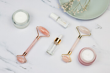 Face Massage pink quartz roller, face creams, face serum oil and flowers on white marble background. Skin care facial massage and relaxation. Anti-aging and lifting concept at home.