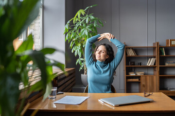 Smiling young female employee relaxing at workplace, sitting in chair stretching arms, doing...