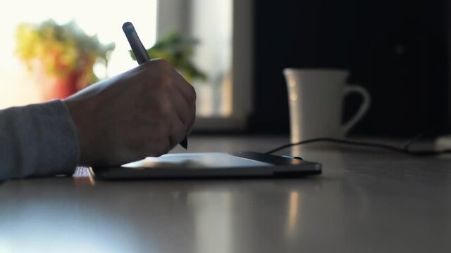 Close-up of designer, illustrator using graphics tablet and pen while sitting at table indoors in morning. Male artist's hand drawing with stylus, retouching, working on professional tablet, side view