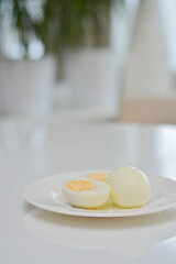Boiled eggs on a kitchen table, healthy breakfast and protein rich diet. Eggs on a plate with a knife on a side, airy and white background with green plants and a light reflection from the table.