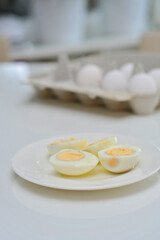 Boiled eggs on a kitchen table, healthy breakfast and protein rich diet. Eggs on a plate with a knife on a side, airy and white background with green plants and a light reflection from the table.