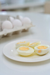 Boiled eggs on a kitchen table, healthy breakfast and protein rich diet. Eggs on a plate with a knife on a side, airy and white background with green plants and a light reflection from the table.