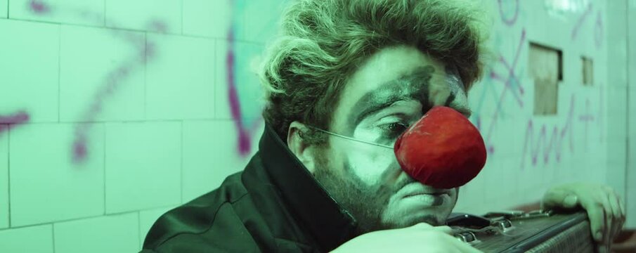 Sad clown sitting with suitcase in subway and looking at train passing by him