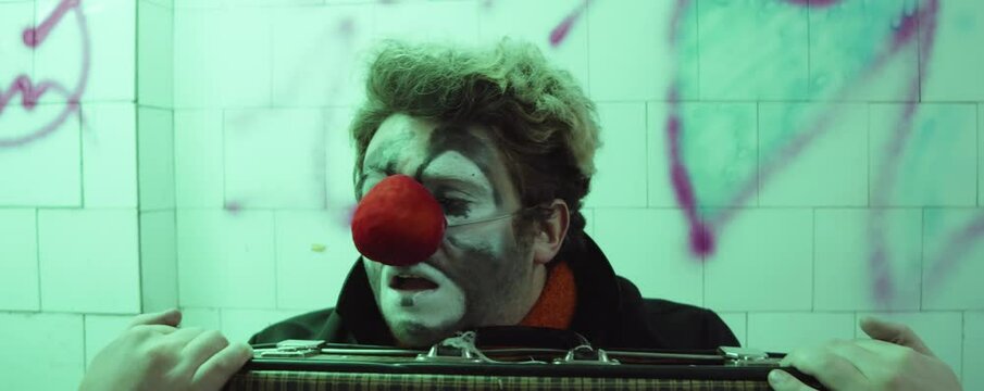 Unhappy clown sitting with suitcase in subway and waiting for train