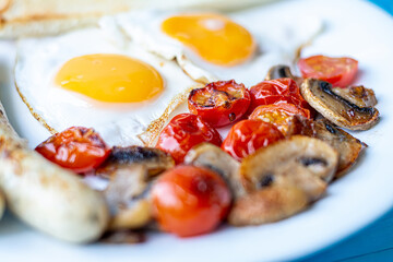 Breakfast of fried eggs, grilled sausages, fried mushrooms and cherry tomatoes macro close-up on a white plate.