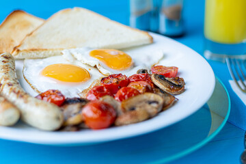 Breakfast of fried eggs, grilled sausages, fried mushrooms and cherry tomatoes and crispy toast on a white plate on a wooden blue table with a glass of orange juice.