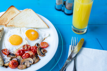 Breakfast of fried eggs, grilled sausages, fried mushrooms and cherry tomatoes and crispy toast on a white plate on a wooden blue table with a glass of orange juice.