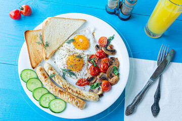 Breakfast of fried eggs, grilled sausages, fried mushrooms and cherry tomatoes, fresh cucumber slices and crispy toast on a white plate on a wooden blue table with a glass of orange juice. Top view.