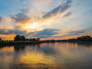 Fototapeta premium Evening sky with dramatic clouds over the river. Dramatic sunset in clouds and river. Nature landscape, reflection, blue cloudly sky and orange sunlight, landscape during sunset.