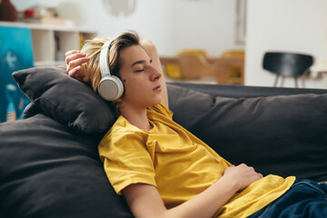 teenager boy listening music on headphones and relaxing on sofa at home