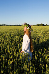 Long haired girl dreesed in white with the frower wreath on the head goes through the corn field in the midsummer day. Blue sky and green meadow