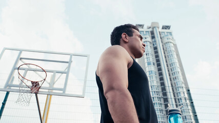 low angle view of sportive mixed race man with sports bottle near basketball ring outdoors.