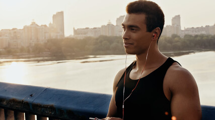 smiling bi-racial sportsman in earphones listening music on city bridge over river.