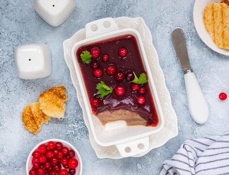 Cold appetizer, tender chicken liver pate with cranberry jelly garnished with cranberries and parsley in a white dish on a light concrete background. Liver recipes.
