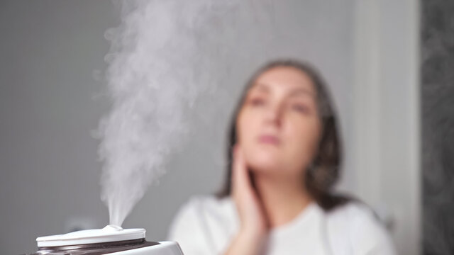 Young brunette woman applies moisturizing cream on dry face skin sitting in room near humidifier vaporing flow of steam into air closeup.