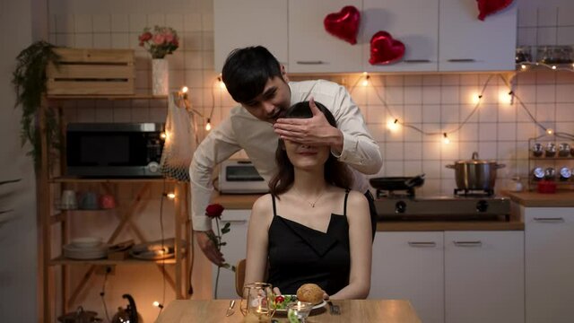 asian male wearing suit covering girlfriend’s eyes and surprising her with a red rose while having romantic valentine’s day dinner at home. woman kissing boyfriend as she receives flower