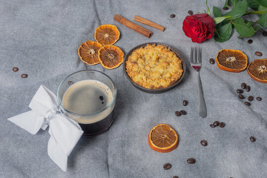 Cup with black coffee, with coffee beans, dried oranges, apple pie, cinnamon stick and red rose on a brown background.