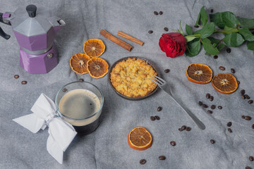 Cup with black coffee, Coffee pot with coffee beans, dried oranges, apple pie, cinnamon stick and red rose on a brown background.