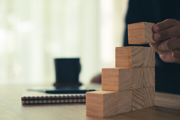 Business man wearing black shirt sitting at desk with coffee and notebook hand stacking wooden blocks for in shape of staircase. Business development, strategy concept. Building success foundation.