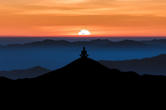Silhouette of young male sitting practices yoga and meditating in lotus position alone on top of the mountain with beautiful sunrise and bird over the sky. He felt calm and happy.