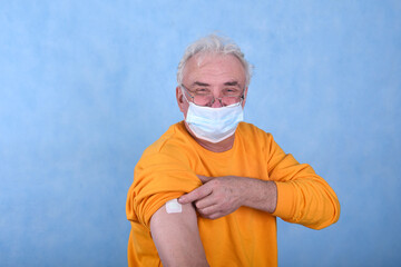 A gray-haired man of retirement age demonstrates the injection site of the vaccine on his arm on a blue background with a joyful and cheerful expression on his face