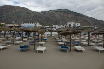 Fototapeta na wymiar evocative image of a sandy beach with empty sunbeds and straw umbrellas with mountains and clouds in the background