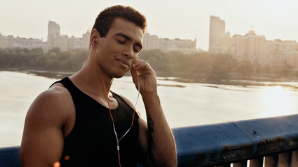 smiling mixed race man with closed eyes listening music in earphones on bridge over river.
