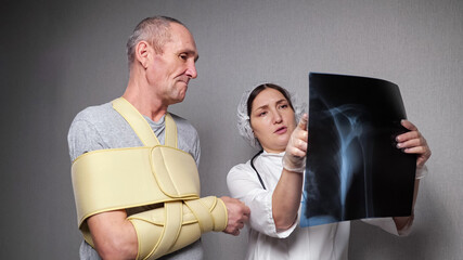 Lady orthopedist shows problems with joints on x-ray photograph to elderly man patient with fixating bandage over torso at grey wall closeup.