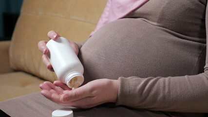 Muslim woman wearing pink hijab and dress caresses softly pregnant belly and drinks vitamins...