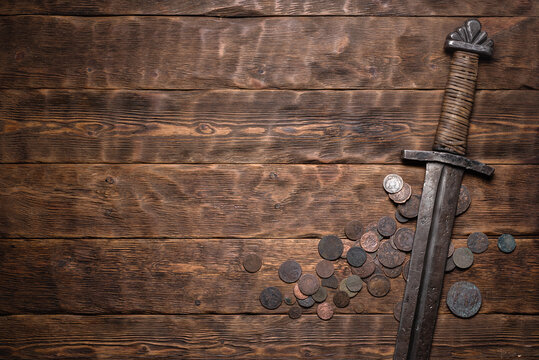 Knight sword and old coins on the wooden flat lay table background with copy space.