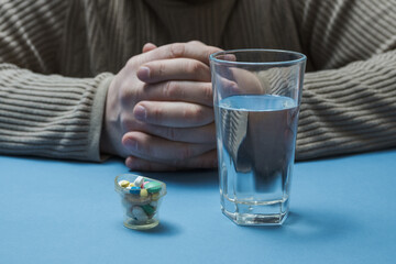 A man is sitting at a table with a glass of water and a medical cup with medicines in front of him.
