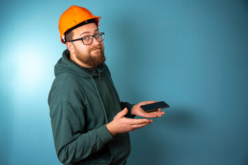 Cute Caucasian bearded construction worker with safety helmet on head and smartphone in hand. Engineer in orange helmet isolated on background. Construction worker concept .