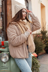 Stylish beautiful young woman in fashionable outerwear with a knitted hat, beige down jacket with a sweater and a scarf stands near a rectro green scooter in the city near a cafe. Winter female style