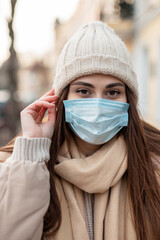 Modern portrait of a young pretty woman with a medical protective mask in fashionable winter clothes with a knitted hat, jacket and scarf walks in the city and looks at the camera. COVID-19 style