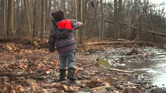 A toddler boy throws a rock into a river on a cold, autumn day, slow motion.