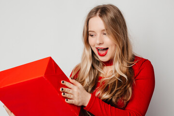 Portrait of a surprised young woman in a red sweater trying to guess what inside gift box, shaking present and looking amazed, standing over white background. Valentine's Day present concept