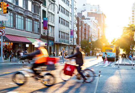 Delivery men on bikes riding through a busy intersection on 14th Street in New York City with sunset in the background