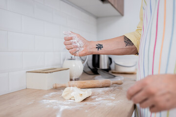 Cropped view of tattooed man pouring flour on dough near rolling pin on worktop in kitchen.