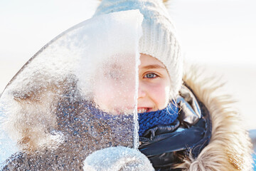 Portrait of smiling little girl looks through a transparent piece of ice on a frosty winter day