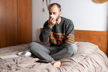 Man with headphones, mobile phone and face mask, sitting on his bed and confined to his room
