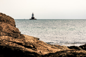 Lighthouse in ocean near rocky seaside on cloudy day in New England.