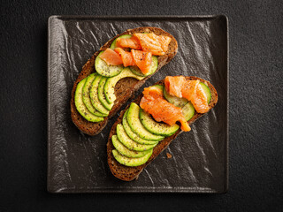 Avocado toast with salmon and cucumbers on dark background top view
