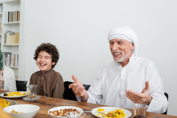 happy muslim man pointing with hands near laughing arabian grandson during family dinner.