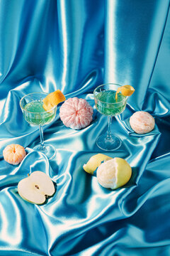 Creative modern still life made against blue satin with exotic drinks in champagne glasses, peeled citrus fruits and apple. Retro minimal background.