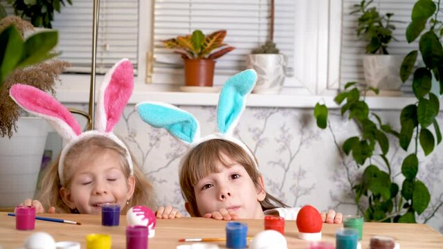 a little girl and a boy with Easter bunny ears jump out from under the table and laugh, chicken eggs and paint brushes are on the table. the concept of a religious holiday