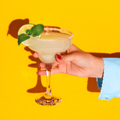 Female hand holding glass with margarita cocktail isolated on bright yellow neon background with...