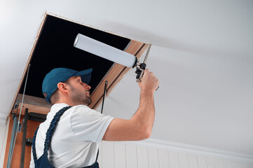 a uniformed specialist fills the gap between the door frame of the attic staircase and the mounting box with polyurethane foam. - obrazy, fototapety, plakaty