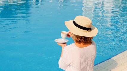 The culture of drinking tea and coffee. Woman with straw hat drinking hot drink from a cup near blue swimming pool, luxury good morning, start of the day