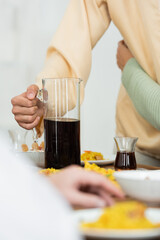 cropped view of man holding jug with tea during family dinner on blurred foreground.