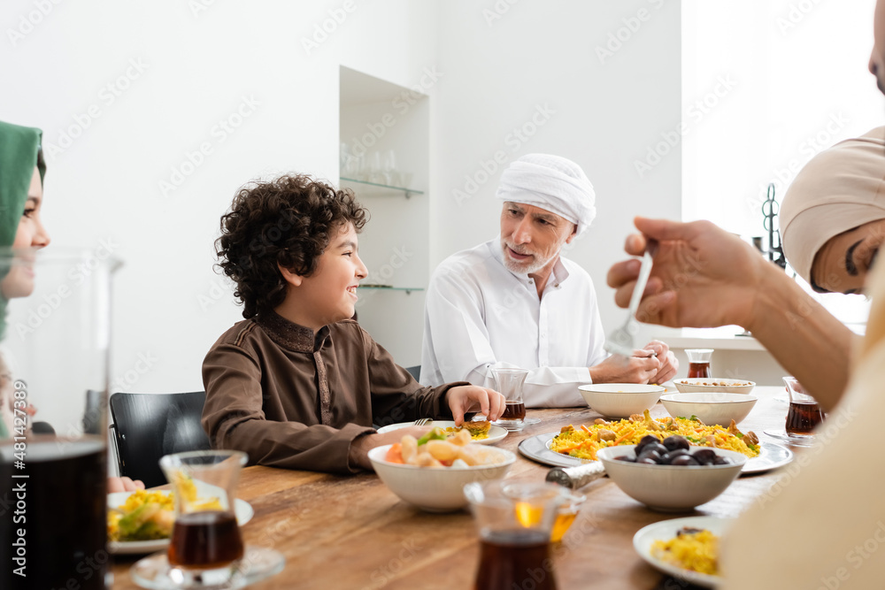 Wall mural muslim man having dinner with happy multiethnic grandchildren and blurred family.